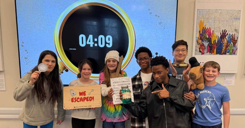 Middle school kids at their library holding up a sign and items from completing their sherlock holms escape game.