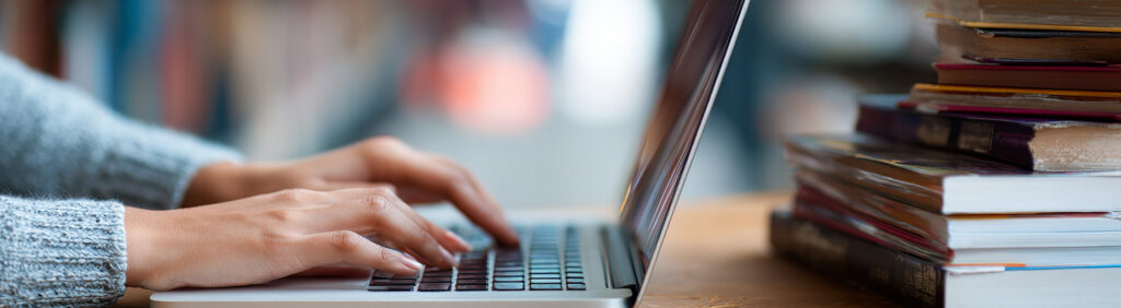 Lady at the library on her computer with a stack of books on the right of the image.